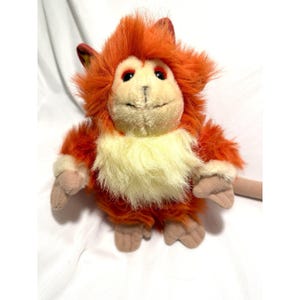 May include: A plush toy monkey with orange fur, a cream-colored chest, and tan hands and feet. The monkey has a smiling expression and is set against a white background. The toy has a long, tan tail.