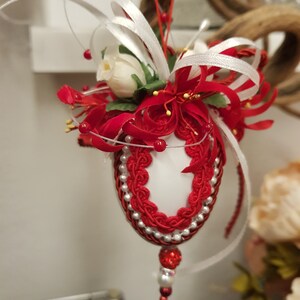 Può includere: Un ornamento bianco a forma di uovo decorato con pizzo rosso e perline bianche. L'ornamento è sormontato da un nastro bianco e un fiore rosso.