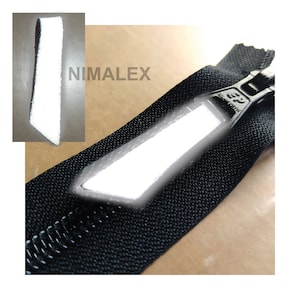 Puede incluir: Un tirador de cremallera reflectante blanco con una cinta de cremallera negra. El tirador de cremallera está unido a una cremallera negra. El tirador de cremallera tiene forma rectangular y tiene una superficie reflectante. La cinta de cremallera está hecha de una tela tejida. El tirador de cremallera mide aproximadamente 5 cm de largo y 2,5 cm de ancho.