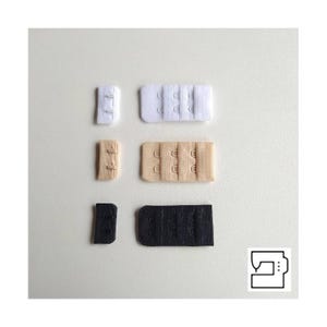 Op de afbeelding: Een assortiment bh-haak- en oogsluitingen in wit, beige en zwart. De sluitingen variëren in grootte en aantal haken en ogen, geschikt voor naaien en kledingconstructie. Een naaimachine-icoon staat in de rechter benedenhoek.