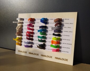 May include: A rectangular wooden display board featuring a color chart of thirty-six small fabric elastic loops arranged in four columns. Each loop is labeled with a number and color name, with the text NIMALEX.DE printed along the bottom edge.