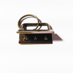 Op de afbeelding: Een gouden metalen schuifgesp met drie tanden en een bevestigde ring. De gesp is ontworpen om te worden gebruikt voor het bevestigen van banden of riemen.