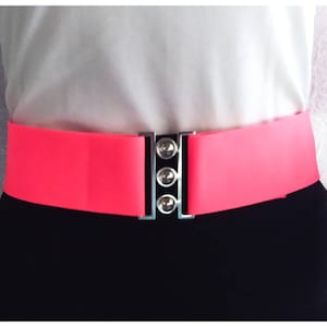 Könnte beinhalten: Ein leuchtend pinkfarbener Gummibandgürtel mit einer silbernen Schnalle mit drei runden Knöpfen. Der Gürtel wird über einem weißen Hemd und einer schwarzen Hose getragen.