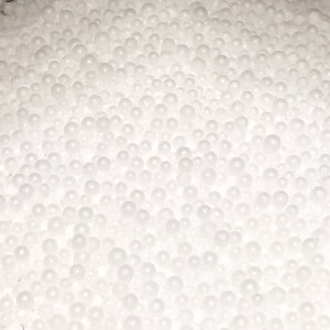 Può includere: Primo piano di una massa di piccole perle bianche e sferiche. Le perle variano in dimensioni e sono fittamente impacchettate, creando un aspetto strutturato e a bolle. L'illuminazione è morbida, evidenziando le forme arrotondate.