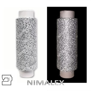 Könnte beinhalten: Eine Spule silbernen Metallic-Garn zum Nähen. Das Garn ist um eine weiße Kunststoffspule mit einem weißen Etikett gewickelt. Die Spule liegt auf einer weißen Oberfläche.