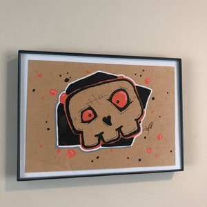 May include: A framed illustration of a cartoon skull with red eyes and a black background. The skull is drawn in a simple, cartoon style and has a friendly expression. The background is a light brown color with black and orange splatters.