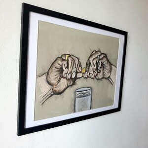 May include: A framed drawing of two hands holding a small yellow object, with the word "SAME" spelled out on the fingers of one hand. A glass of water is in the background.