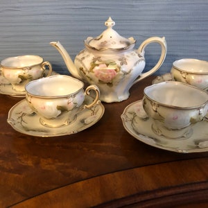May include: A vintage porcelain tea set featuring a teapot and four teacups with saucers. The set is decorated with pink roses, green leaves, and gold trim. The teapot has a lid and a curved spout. The teacups and saucers are delicately painted.