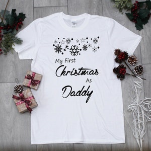 May include: White t-shirt with black text that reads "My First Christmas As Daddy" with a row of snowflakes above the text.