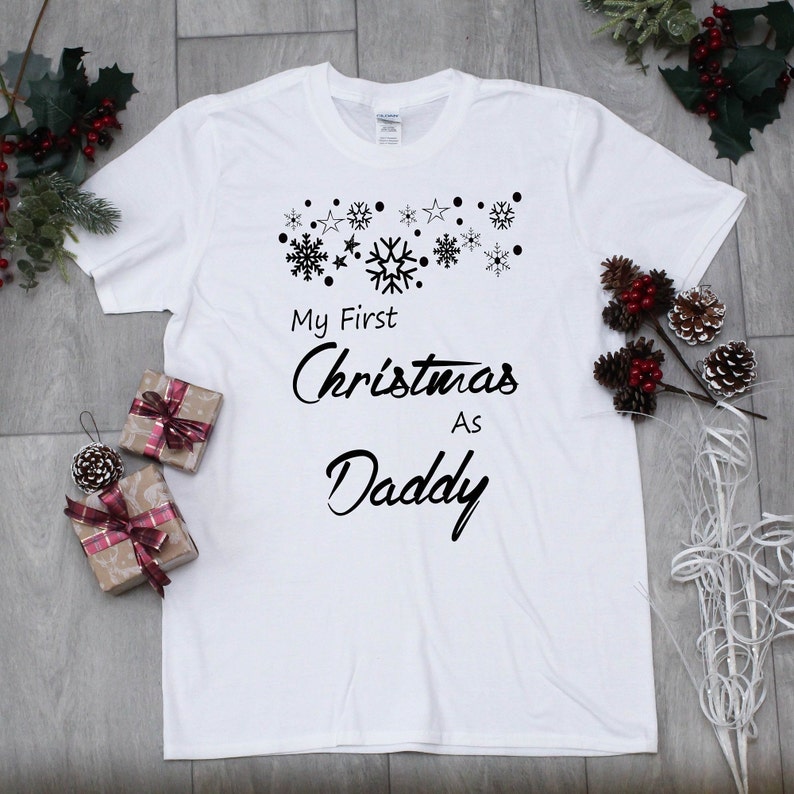 May include: White t-shirt with black text that reads "My First Christmas As Daddy" with a row of snowflakes above the text.