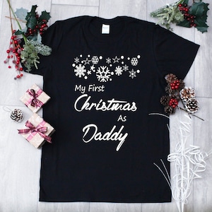 May include: Black t-shirt with white snowflakes and the text "My First Christmas As Daddy".