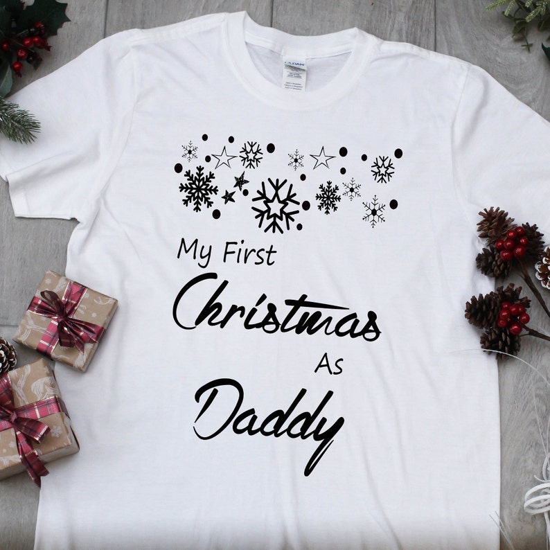 May include: White t-shirt with black text that reads "My First Christmas As Daddy" with a design of snowflakes and stars above the text.