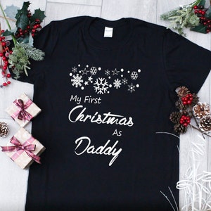 May include: Black t-shirt with white text that reads "My First Christmas As Daddy" with snowflakes and stars above the text.