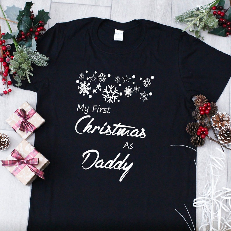 May include: Black t-shirt with white text that reads "My First Christmas As Daddy" with snowflakes and stars above the text.
