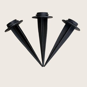 May include: Three black plastic ground stakes arranged in a fan shape. Each stake features a pointed bottom, a ribbed vertical shaft, and a flat circular top flange designed for securing items into the ground with a sturdy, matte finish.
