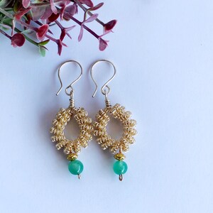 Può includere: Orecchini in filo d'oro con gocce di pietre preziose verdi. Gli orecchini sono realizzati in filo d'oro intrecciato in una forma circolare. Le pietre preziose sono di colore verde chiaro e sono attaccate alla parte inferiore degli orecchini.