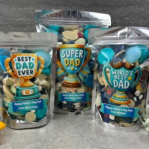 May include: Three clear cellophane bags filled with colorful candies. Each bag features a trophy design with text: "BEST DAD EVER", "SUPER DAD", and "WORLD'S BEST DAD". The bags are labeled "Happy Father's Day Dad" with names.