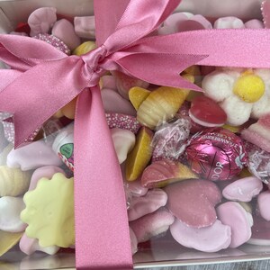 K&ouml;nnte beinhalten: Eine transparente Box gef&uuml;llt mit rosa und wei&szlig;en S&uuml;&szlig;igkeiten, gebunden mit einem rosa Satinband. Die S&uuml;&szlig;igkeiten umfassen Herzen, Blumen und andere Formen.