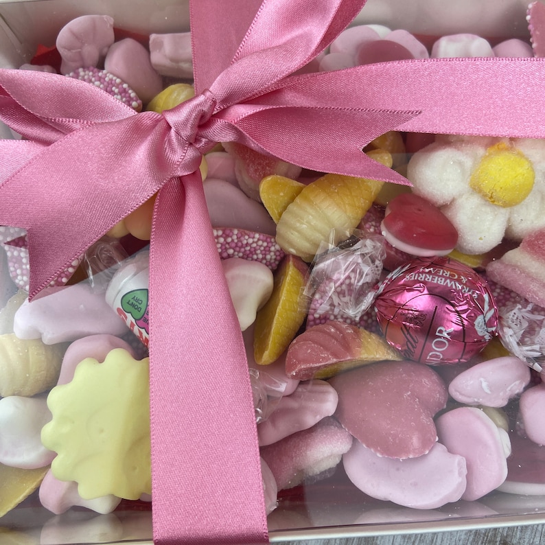 K&ouml;nnte beinhalten: Eine transparente Box gef&uuml;llt mit rosa und wei&szlig;en S&uuml;&szlig;igkeiten, gebunden mit einem rosa Satinband. Die S&uuml;&szlig;igkeiten umfassen Herzen, Blumen und andere Formen.
