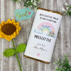 May include: A chocolate bar wrapper with gold edging, displaying a rainbow design and the text "JUST A LITTLE TREAT TO SAY A VERY BIG thank you MISS LITTLE Love from Ava Mae x". A crocheted yellow sunflower and a "Good" card are also present.