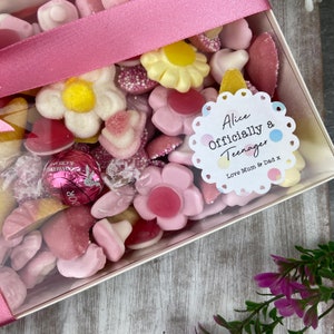 K&ouml;nnte beinhalten: Eine transparente Box gef&uuml;llt mit rosa und wei&szlig;en S&uuml;&szlig;igkeiten, darunter Gummib&auml;rchen in Form von Blumen, Herzen und anderen Formen. Ein wei&szlig;er Aufkleber mit einem rosa gewellten Rand lautet "Alice Offiziell ein Teenager Liebe Mama & Papa x".
