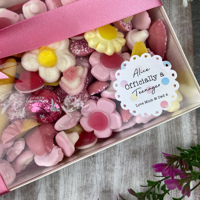 K&ouml;nnte beinhalten: Eine transparente Box gef&uuml;llt mit rosa und wei&szlig;en S&uuml;&szlig;igkeiten, darunter Gummib&auml;rchen in Form von Blumen, Herzen und anderen Formen. Ein wei&szlig;er Aufkleber mit einem rosa gewellten Rand lautet "Alice Offiziell ein Teenager Liebe Mama & Papa x".