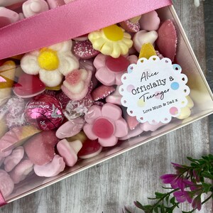 K&ouml;nnte beinhalten: Eine transparente Box gef&uuml;llt mit rosa, wei&szlig;en und gelben S&uuml;&szlig;igkeiten. Ein wei&szlig;er runder Aufkleber mit einem rosa, blauen und gelben Tupfenmuster lautet "Alice Offiziell ein Teenager Liebe Mama & Papa x".