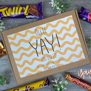 Pode incluir: Uma caixa de cartão castanho com um cartão branco no seu interior. O cartão tem um padrão ondulado amarelo e branco e diz "OLLIE YAY! YOU DID IT! WELL DONE ON PASSING YOUR EXAMS LOVE MUM AND DAD X". A caixa é rodeada por barras de chocolate.