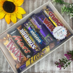 K&ouml;nnte beinhalten: Eine wei&szlig;e Geschenkbox gef&uuml;llt mit einer Auswahl an Schokoladenriegeln, darunter Twix, Mars, Starbar, Boost, Flake, Wispa und Crunch. Die Box hat einen durchsichtigen Deckel und ein Etikett mit der Aufschrift "Happy Birthday Lucy" in rosa und blau.