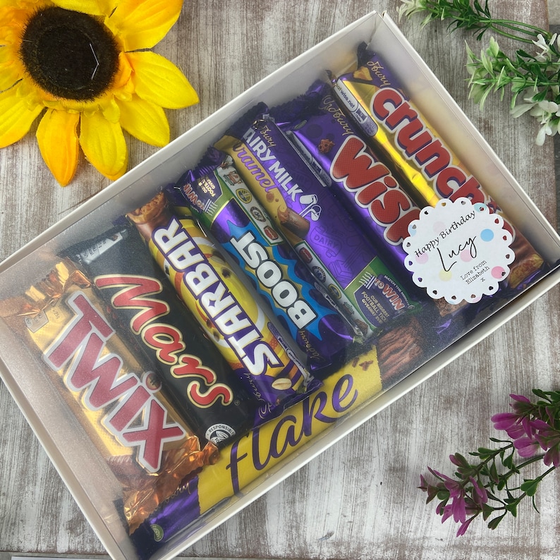 K&ouml;nnte beinhalten: Eine wei&szlig;e Geschenkbox gef&uuml;llt mit einer Auswahl an Schokoladenriegeln, darunter Twix, Mars, Starbar, Boost, Flake, Wispa und Crunch. Die Box hat einen durchsichtigen Deckel und ein Etikett mit der Aufschrift "Happy Birthday Lucy" in rosa und blau.