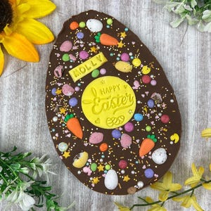 May include: A large, chocolate Easter egg decorated with colorful sprinkles, candies, and carrot-shaped candies. The egg has a yellow circle with "Happy Easter" and a name "Molly" on it. The egg is on a light wood surface.
