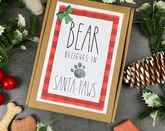 May include: A brown cardboard box with a red and white plaid border and the text "BEAR BELIEVES IN SANTA PAWS". A green bow is in the upper left corner. The box is surrounded by dog treats, holly, and a small white bear.