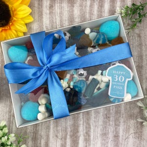 Könnte beinhalten: Eine transparente Box gefüllt mit verschiedenen Süßigkeiten, gebunden mit einem blauen Band. Die Süßigkeiten umfassen blaue, weiße und braune Süßigkeiten, mit fußballförmigen Süßigkeiten. Ein Etikett lautet "Happy 30th Birthday Paul."