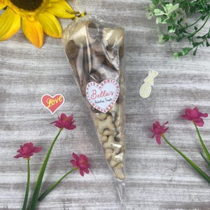May include: A clear plastic cone filled with dog treats. The cone has a heart-shaped label that says "Bella's Valentine Treats". There are also two small heart-shaped stickers, one that says "Love" and one that says "XO".