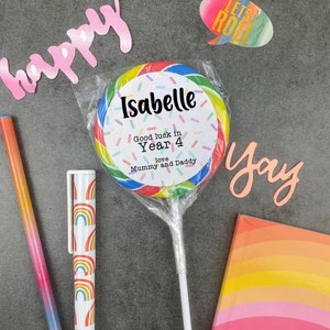 May include: A colorful lollipop with a personalized label that reads "Isabelle, Good luck in Year 4, love Mummy and Daddy." The lollipop is wrapped in clear cellophane. Other items include a pen, a notebook, and cut-out words.