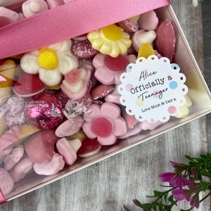 K&ouml;nnte beinhalten: Eine transparente Geschenkbox gef&uuml;llt mit rosa, wei&szlig;en und gelben S&uuml;&szlig;igkeiten. Ein wei&szlig;er runder Aufkleber mit einem rosa, blauen und gelben Tupfenmuster lautet "Alice Offiziell ein Teenager Liebe Mama & Papa x".