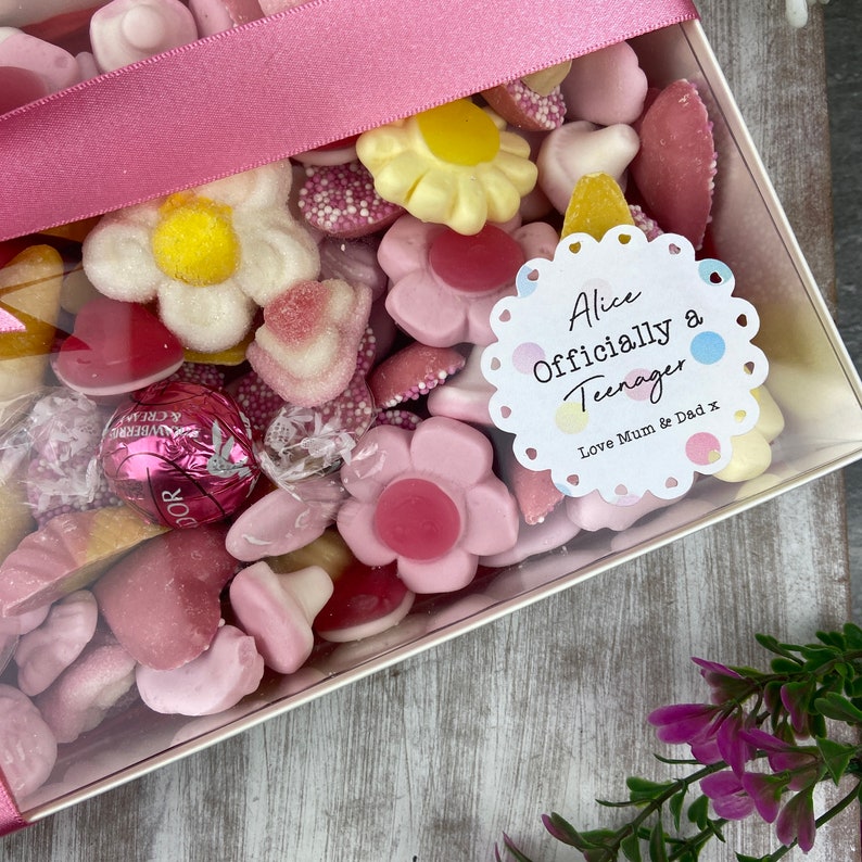 K&ouml;nnte beinhalten: Eine transparente Geschenkbox gef&uuml;llt mit rosa, wei&szlig;en und gelben S&uuml;&szlig;igkeiten. Ein wei&szlig;er runder Aufkleber mit einem rosa, blauen und gelben Tupfenmuster lautet "Alice Offiziell ein Teenager Liebe Mama & Papa x".