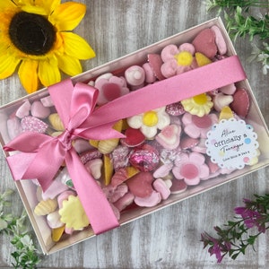 K&ouml;nnte beinhalten: Eine durchsichtige Box gef&uuml;llt mit rosa und gelben S&uuml;&szlig;igkeiten, die mit einem rosa Band gebunden ist. Die Box hat einen Aufkleber mit der Aufschrift "Alice Offiziell ein Teenager" mit dem Text "Love Mum & Dad x".