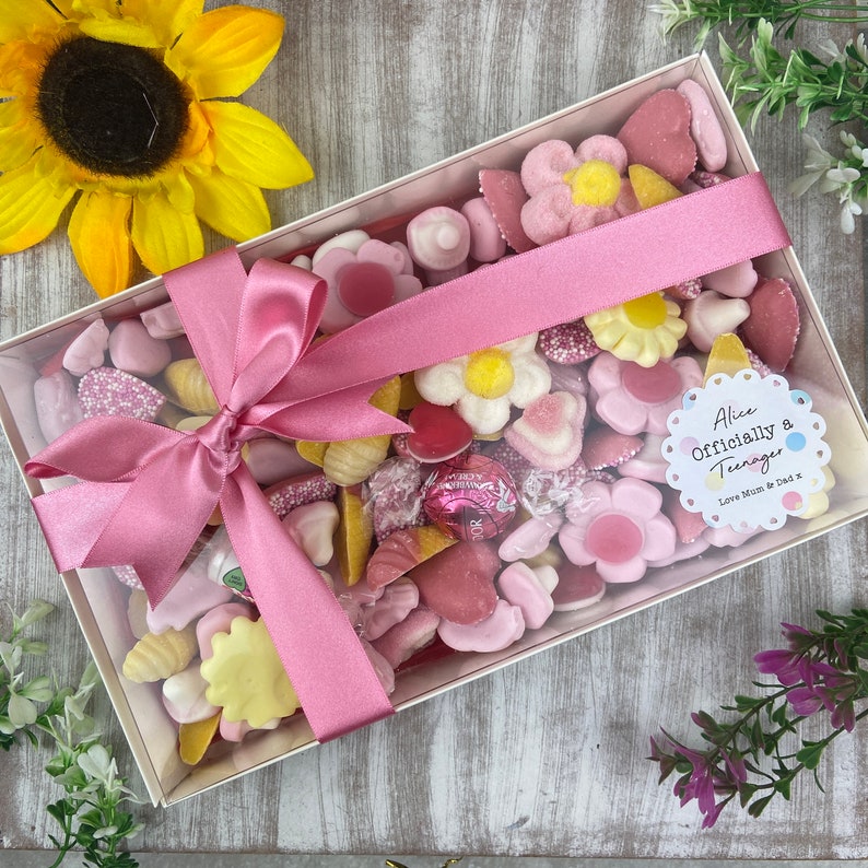 K&ouml;nnte beinhalten: Eine durchsichtige Schachtel gef&uuml;llt mit rosa und gelben S&uuml;&szlig;igkeiten, die mit einem rosa Band gebunden ist. Die Schachtel hat einen Aufkleber mit der Aufschrift "Alice Officially a Teenager Love Mum & Dad x".