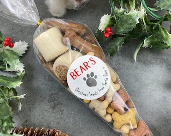 May include: A clear cellophane bag filled with assorted dog treats, including bone-shaped and square biscuits. The bag is sealed with a gold twist tie and features a circular sticker that reads "Bear's Christmas Treats From Santa Paws".