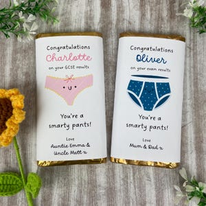 Peut inclure: Deux barres de chocolat avec un emballage en feuille d'or. L'une présente un motif de sous-vêtements roses et le nom "Charlotte" avec le texte "Félicitations pour vos résultats au GCSE". L'autre a un motif de sous-vêtements bleus et le nom "Oliver" avec le texte "Félicitations pour vos résultats d'examen".