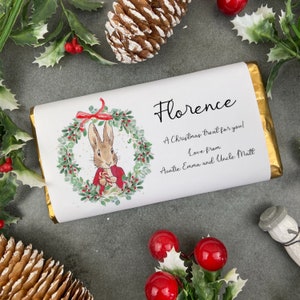 以下が含まれることがあります： 金色のフォイルで包まれたチョコレートバー。ラベルには、クリスマスの花輪の中のウサギ、名前「フローレンス」、休日のメッセージが描かれています。パーソナライズされたチョコレートバーです。