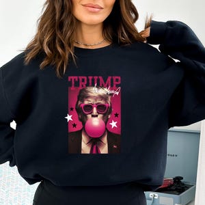 May include: Black sweatshirt with a graphic print of a man wearing sunglasses and blowing a pink bubble. The graphic includes the text "TRUMP" and a signature.