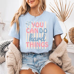 May include: A light blue short-sleeve t-shirt featuring a graphic design with vertical beige stripes and a small blue bow. The text reads "YOU CAN DO hard THINGS" in a mix of pink and blue lettering, centered on the front of the garment.