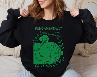 Puede incluir: Sudadera negra con un gráfico verde de una persona sosteniendo un globo terráqueo. El texto dice "Fundamentalmente y ambientalmente amigable".