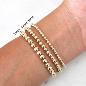 May include: Three gold beaded bracelets, with bead sizes of 3mm, 4mm, and 5mm, are displayed on a wrist. The bracelets are shiny and round, with the sizes labeled in the image.