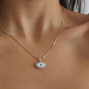 Könnte beinhalten: Eine goldene Kettenkette mit einem weißen und blauen bösen Augen-Anhänger. Der Anhänger ist von kleinen, klaren Steinen umgeben.