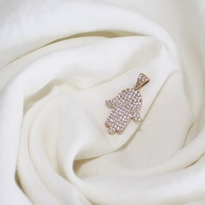 Könnte beinhalten: Ein goldener Hamsa-Hand-Anhänger mit klaren, funkelnden Steinen.