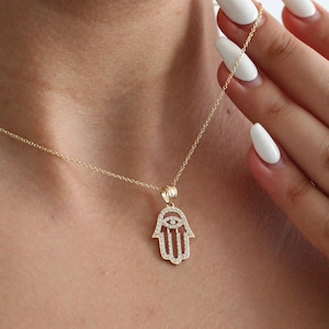 Könnte beinhalten: Eine goldene Halskette mit einem Hamsa-Hand-Anhänger, der mit kleinen, klaren Steinen besetzt ist. Die Hamsa-Hand hat ein böses Auge in der Mitte.