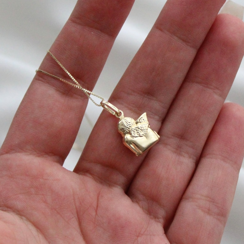 May include: A gold chain necklace with a small angel pendant. The angel is facing to the right and has its wings spread out.